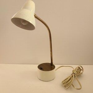 May include: A vintage white desk lamp with a flexible brass neck and a round base. The lamp has a classic design with a white lampshade and a cord. The lamp is approximately 12 inches tall.