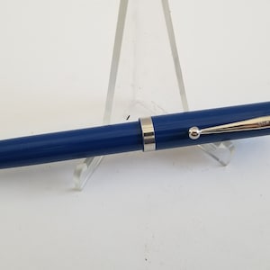 May include: A blue fountain pen with a silver clip and trim. The pen is displayed on a clear acrylic stand against a white background. The pen's design is simple and elegant.