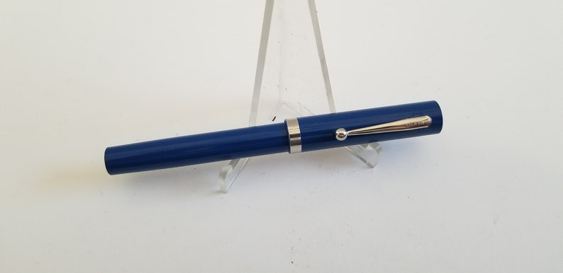 May include: A blue fountain pen with a silver clip and trim. The pen is displayed on a clear acrylic stand against a white background. The pen's design is simple and elegant.