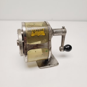 May include: Vintage Giant pencil sharpener with a clear plastic casing and a silver-colored metal frame. The sharpener has a crank handle with a black knob and a label that reads "Giant Made by Automatic Pencil Sharpener Co. Chicago, U.S.A."