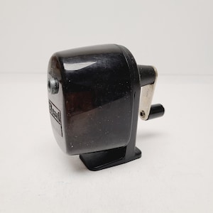 May include: A black pencil sharpener with a silver crank and a small black handle. The sharpener has a rectangular base and a rounded top. The brand name "Bassett" is visible on the side.