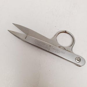 May include: Stainless steel thread clipper with the text "COMPTON-U-SET THREAD CLIPPER Stainless Steel U.S.A." The clipper has a ring handle and pointed blades. The tool is designed for cutting threads and is likely used in sewing or crafting.