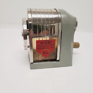 May include: A vintage Apsco Deluxe Giant automatic pencil sharpener with a grey metal base, a chrome-plated top, and a clear plastic shavings receptacle. A red shield-shaped label on the front reads: DELUXE GIANT AUTOMATIC PENCIL SHARPENER CO. ROCKFORD, ILL., U.S.A. Apsco.