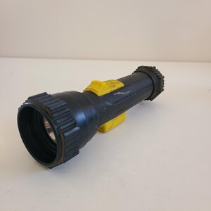 May include: A black and yellow flashlight with a ribbed head and a textured end. The flashlight has a yellow switch and a yellow base. The flashlight is lying on a white surface.