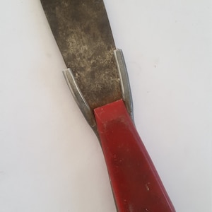May include: A red-handled putty knife with a metal blade. The blade is dirty and has a worn appearance.
