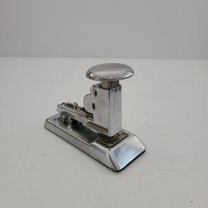 May include: A silver metal stapler with a round, silver top. The stapler has a base and a lever on the side.