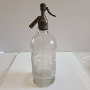 May include: A vintage glass bottle with a metal siphon top. The bottle is clear and has a white etched label that reads "Schweppes Limited London Special Aerated Water."