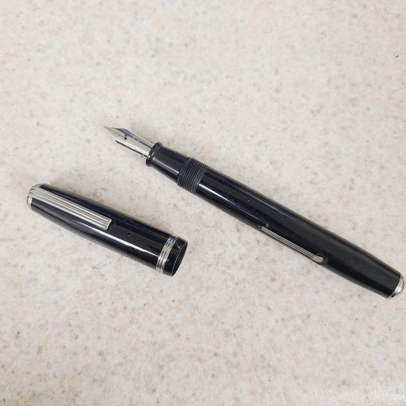 May include: A black fountain pen with a silver nib and accents. The pen's cap is detached, revealing the pen's silver trim. The pen is lying on a light-colored surface. A classic writing instrument.