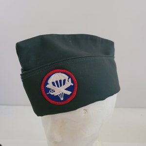 May include: A dark green military-style hat with a red, white, and blue patch featuring a parachute and airplane. The patch is centered on the front of the hat.