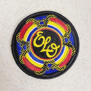 May include: Embroidered patch featuring the letters "ELO" in a stylized script within a black circle. The design includes concentric rings of blue, yellow, and red, with decorative elements in gold and black. The patch has a black border.