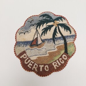 May include: A circular punch needle textile patch with a scalloped edge featuring a tropical beach scene with a palm tree, a sailboat on the water, and birds in the sky. The text PUERTO RICO is embroidered in white along the bottom brown border.
