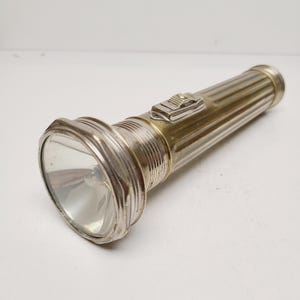 May include: A vintage flashlight with a silver and gold-toned metal body. The flashlight has a ribbed design on the cylindrical portion and a rectangular switch. The lens is clear, and the light is off.