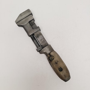 May include: A vintage adjustable wrench featuring a weathered metal head and a threaded adjustment screw. The handle is constructed from light-colored wood, secured with two dark metal rivets, showing signs of age and heavy use throughout the tool.