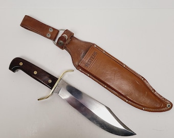 May include: A large hunting knife with a polished steel blade and a dark wooden handle. The knife has a brass guard and is accompanied by a brown leather sheath. The sheath is stamped with the word "WESTERN".