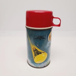 May include: Vintage Thermos brand vacuum flask with a red plastic cup and handle. The cylindrical flask features a blue background with a yellow and white rocket ship design. The word "THERMOS" is printed on the flask.