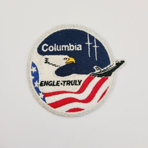 May include: A round, white-bordered patch with a blue background and a white eagle with yellow beak and talons. The eagle is flying over a red, white, and blue American flag. The text "Columbia" is above the eagle, and the text "ENGLE-TRULY" is below the flag.