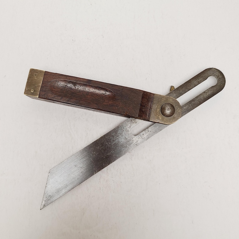 May include: A vintage sliding T-bevel tool featuring a dark brown wooden handle with brass end caps and a pivoting steel blade. The tool includes a central brass locking nut and a slotted metal arm, showing signs of age and wear on the metal surfaces.