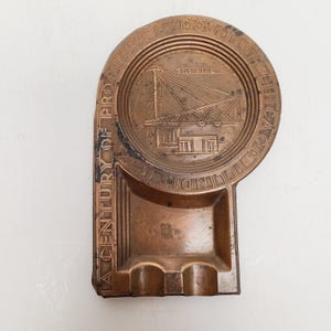 May include: Vintage copper-toned ashtray with a rectangular shape and a circular design. The design features a crane and the words "SKY RIDE" and "A CENTURY OF PROGRESS CHICAGO ILLINOIS EXPOSITION".