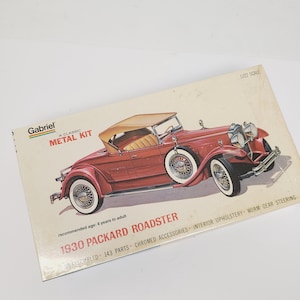 May include: A Gabriel 1/22 scale metal model kit box for a 1930 Packard Roadster. The box features an illustration of a red convertible car with a tan top, wire wheels, and chrome details. Text includes 143 parts, interior upholstery, and worm gear steering.