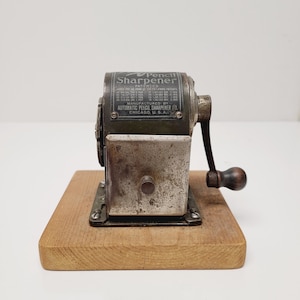 May include: Vintage pencil sharpener with a dark green metal body and a silver-colored base. The sharpener is mounted on a rectangular wooden base. The text on the sharpener reads "Pencil Sharpener" and "Automatic Pencil Sharpener Co. Chicago, U.S.A."