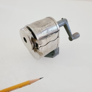 Puede incluir: Un sacapuntas vintage de plata con manivela. El sacapuntas tiene una base gris y el texto "Eagle No. 3 USA" es visible en el lateral.