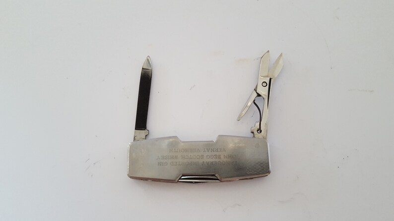 Op de afbeelding: Een zilveren multitool met een mes, een schaar en andere gereedschappen. De tool is gegraveerd met de tekst "AVONDA  PATENTED  USA  MINI  EDC  TOOL  WHITNEY  ILLINOIS".