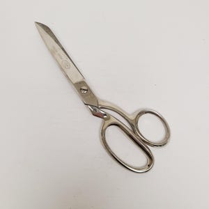 May include: A pair of silver-colored scissors with a curved handle and sharp blades. The scissors have a small oval emblem on the blade. The blades are open, and the scissors are on a white surface.