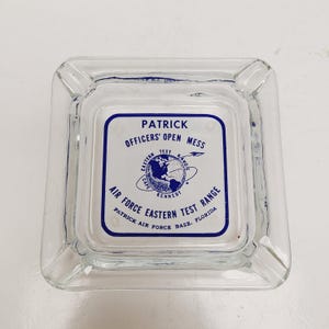 May include: Clear glass ashtray with a square shape and rounded corners. The center features a blue and white graphic with text that reads "PATRICK OFFICERS' OPEN MESS AIR FORCE EASTERN TEST RANGE".