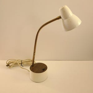 May include: A vintage white desk lamp with a gooseneck arm and a round base. The lamp has a white lampshade and a flexible, brass-colored arm. The base is white with a wooden top and a small switch. A cream-colored power cord extends from the base.