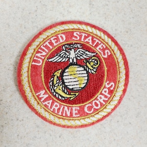 May include: A circular embroidered patch featuring the United States Marine Corps emblem. The patch has a red background with gold lettering and a gold border. The emblem includes an eagle, globe, and anchor.