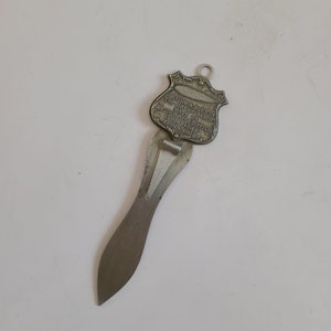 May include: A vintage metal letter opener with a shield-shaped top and a long, curved blade. The shield has text that reads "DURALUMIN USED IN THE AIRSHIP AKRON MADE BY THE GOODYEAR ZEPPELIN CORPORATION AKRON OHIO."