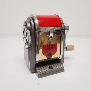 May include: A vintage Dexter Model 6 automatic pencil sharpener with a dark gray metal body, a bright red top cover, and a gold-toned front plate. The front features a red label with black text reading Dexter Model 6 Automatic Pencil Sharpener Company Rockford, Ill. U.S.A. Apsco.
