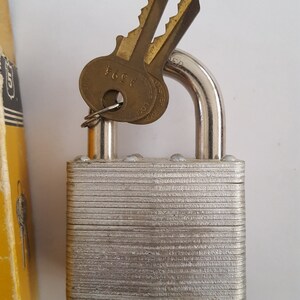 May include: A silver padlock with a brass keyhole and two brass keys. The keys have the text "4564" and "SAFE  LOCK" on them. The padlock has a textured surface.