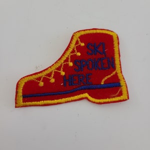 May include: Red felt boot-shaped patch with yellow stitching and blue embroidered text that reads "Ski Spoken Here".