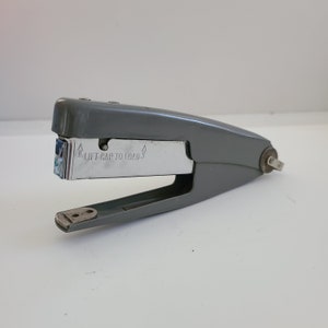 May include: A gray metal stapler with a silver top and a label that reads "Lift cap to load".