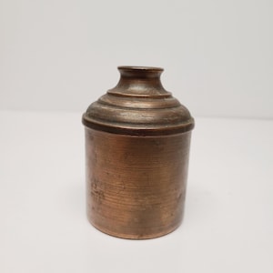 May include: A small, antique copper container with a lid. The container has a cylindrical body and a rounded top with a series of concentric rings. The lid is also rounded and has a small, flared top. The copper has a warm, reddish-brown hue.