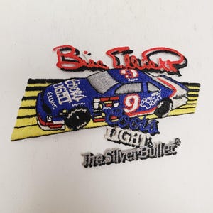 May include: Embroidered patch featuring a blue, white, and red race car with the number 9. The patch includes the words "Big Time Racing", "Coors Light", and "The Silver Bullet". The patch is on a yellow and black background.