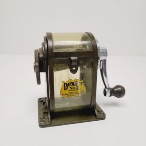 May include: Vintage Dexter No. 3 pencil sharpener. The metal sharpener has a clear plastic top and a crank handle with a wooden knob. The label reads "Dexter No. 3 Improve Made by Automatic Pencil Sharpener Chicago, U.S."