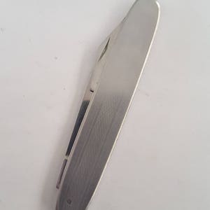 May include: A stainless steel pocket knife with a brushed metal finish. The knife is closed, showing the smooth, rounded top and a small portion of the blade. The clip is visible on the side. The knife is designed for everyday carry.