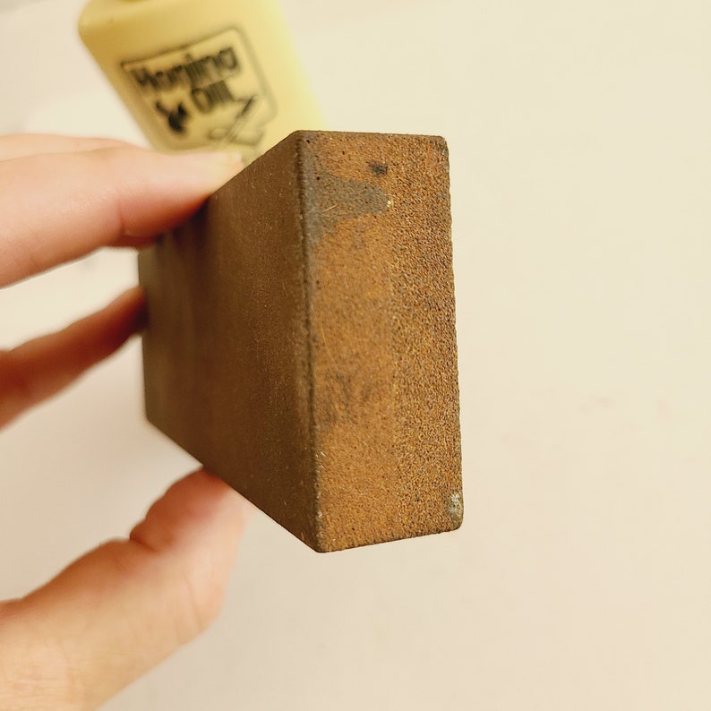 May include: A close-up of a brown sharpening stone with a rough texture. The stone is rectangular and has a slightly rounded edge.