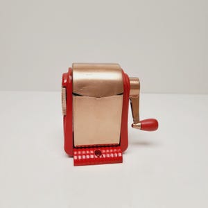 May include: A vintage pencil sharpener with a red base and accents, and a copper-colored body. The sharpener has a hand crank with a red handle. The design is a classic, retro style.