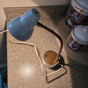May include: A vintage desk lamp with a light blue shade and a flexible, brass-colored neck. The base is white with a wood-grain top and a switch. Two ceramic canisters with rooster and flag designs are in the background.