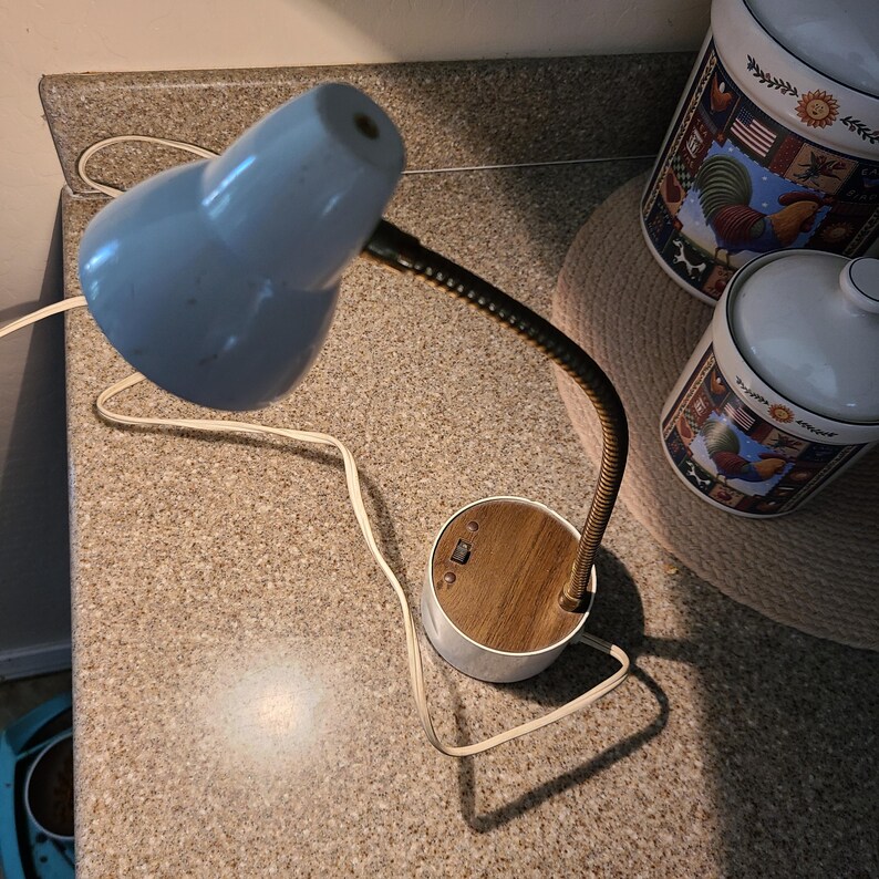 May include: A vintage desk lamp with a light blue shade and a flexible, brass-colored neck. The base is white with a wood-grain top and a switch. Two ceramic canisters with rooster and flag designs are in the background.