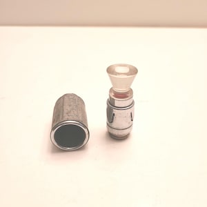 May include: A silver cigarette lighter with a clear glass top and a metal case. The lighter is in two pieces, the top and the bottom. The bottom piece is a silver cylinder with a black interior. The top piece is a clear glass cylinder with a silver rim.