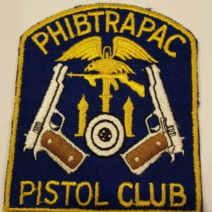 May include: A blue and gold embroidered patch with the text "PHIBTRAPAC PISTOL CLUB". The patch features a stylized eagle with wings spread, holding a rifle and two pistols. The eagle is perched on a target with three arrows.