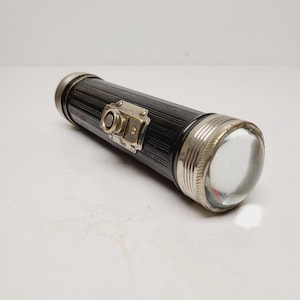 May include: A vintage black flashlight with a ribbed body and silver-toned accents. The flashlight features a clear, rounded lens and a small, decorative metal plate. A classic, functional lighting tool.