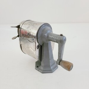 Puede incluir: Un sacapuntas vintage plateado y gris con una manivela de madera. El sacapuntas tiene un cuerpo de metal y una base gris.