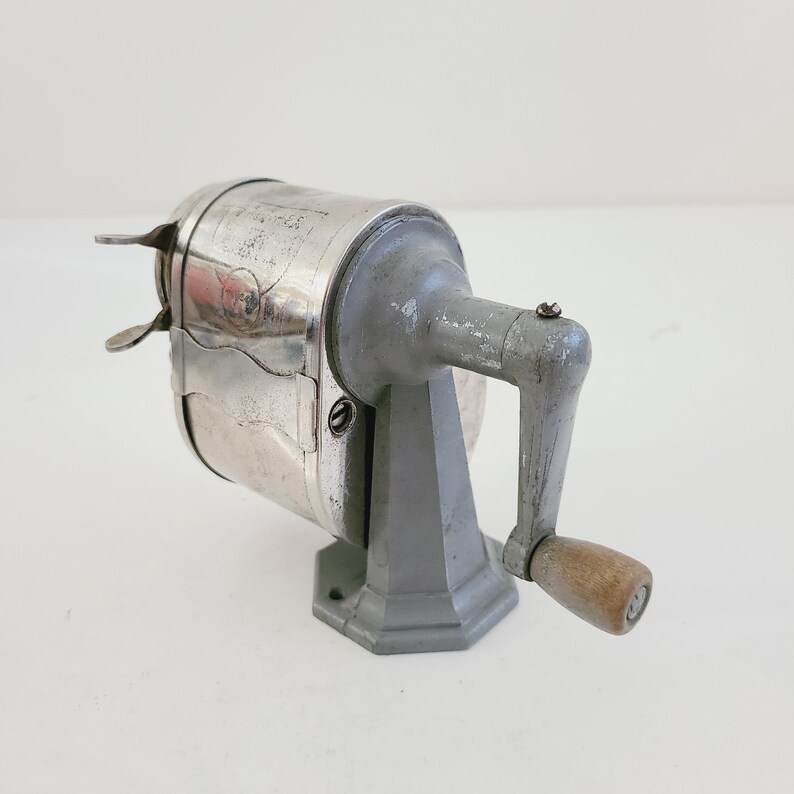 Puede incluir: Un sacapuntas vintage plateado y gris con una manivela de madera. El sacapuntas tiene un cuerpo de metal y una base gris.