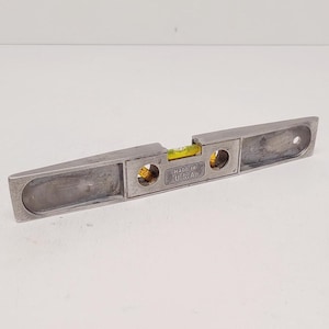 May include: A silver-colored level with a yellow bubble vial. The level has a rectangular shape with rounded ends and two circular openings. The text "MADE IN U.S.A." is visible.