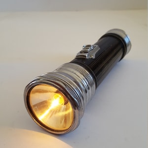 May include: Vintage black and silver flashlight with a bright yellow light emanating from the lens. The flashlight has a ribbed design and a metal switch. A classic lighting tool.
